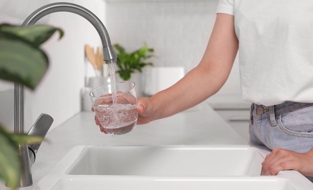 Person Holding Glass Of Water Under Filtered Running Faucet